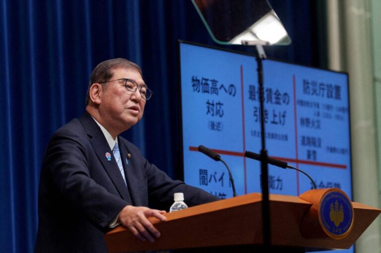 Japanese Prime Minister Shigeru Ishiba addresses the media during a policy briefing in Tokyo, with economic and defense topics displayed behind him.