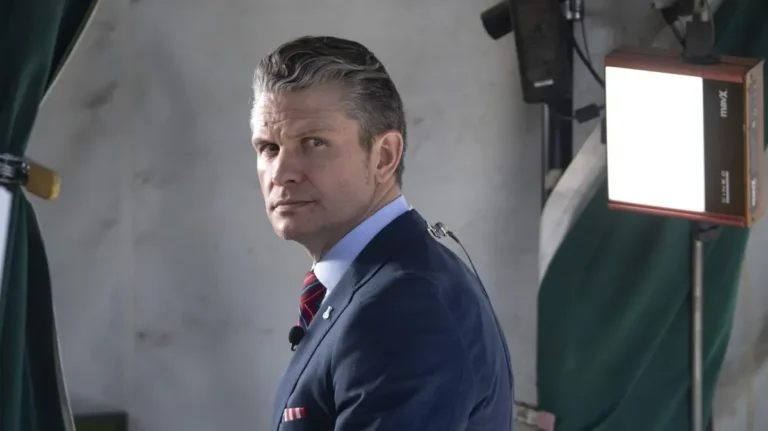 U.S. Defense Secretary Pete Hegseth looks over his shoulder while preparing for a broadcast interview, with studio lights in the background.