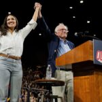 AOC and Bernie Sanders raise their hands together onstage at a packed rally, standing beside a podium with a “Fight Oligarchy” sign during their 2025 national tour.