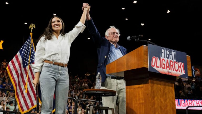 AOC and Bernie Sanders raise their hands together onstage at a packed rally, standing beside a podium with a “Fight Oligarchy” sign during their 2025 national tour.