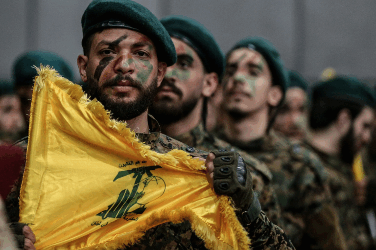 A Hezbollah fighter holds a yellow party flag during a ceremonial formation, with face paint and military uniform.