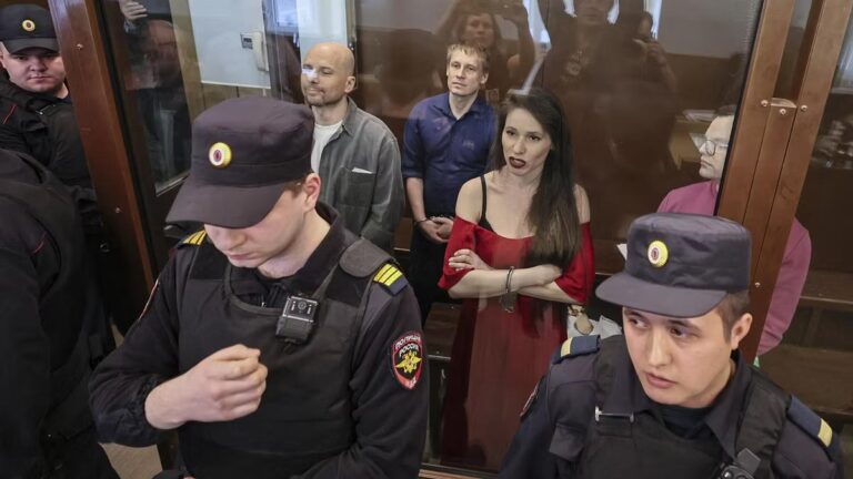 Russian journalists stand in a glass courtroom enclosure as armed guards block the view, following their sentencing for alleged ties to Navalny’s banned group.