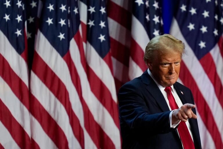 Donald Trump points forward during a speech, standing in front of a backdrop of American flags amid scrutiny over insider trading allegations tied to his tariff policy.