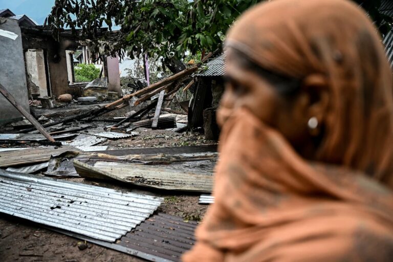 A woman stands near the wreckage of a home destroyed in the Kashmir airport drone attack, as metal debris and rubble cover the ground.